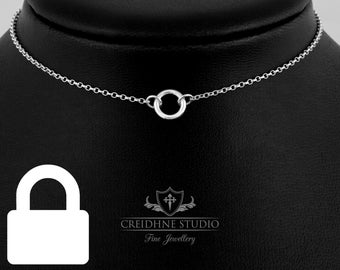Puede incluir: Un collar de cadena de plata con un pequeño colgante circular. El collar está en un expositor negro.