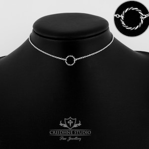 Könnte beinhalten: Eine silberne Choker-Kette mit einem gedrehten Kreis-Anhänger. Die Kette befindet sich auf einer schwarzen Mannequin-Büste.