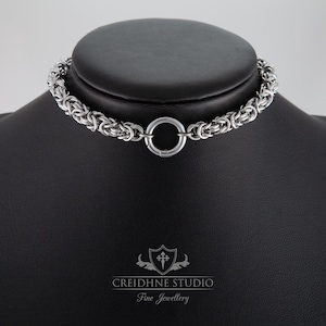 Può includere: Una collana girocollo a maglia d'argento con una grande chiusura circolare. La collana è esposta su una testa di manichino nera. I gioielli sono realizzati con piccoli anelli di metallo intrecciati. Il logo "Creidhne Studio Fine Jewellery" è in basso.