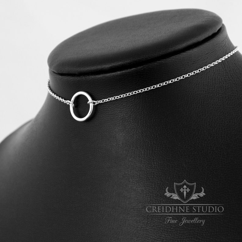 May include: A silver choker necklace with a simple circle pendant. The necklace is on a black display stand.
