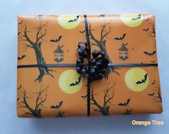 May include: A rectangular gift wrapped in orange paper featuring a repeating pattern of dark brown bare trees, yellow full moons, black bats, and hanging lanterns. The package is tied with a thin black ribbon and topped with a black curly bow.
