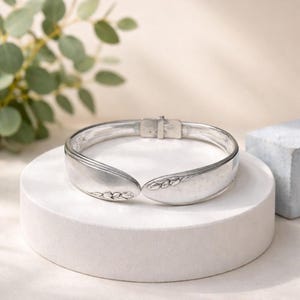 May include: A silver bracelet made from repurposed silverware. The hinged bracelet features a floral design on the spoon ends. The bracelet is displayed on a white pedestal with a neutral background.