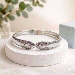 May include: A silver-colored bracelet made from repurposed silverware. The bracelet features a hinged design with decorative floral and fan-like patterns on the spoon ends. The bracelet is displayed on a white, circular platform.