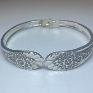 May include: Silver-toned bracelet crafted from repurposed silverware. The bracelet features a hinged clasp and floral detailing on the spoon bowl ends. A unique piece of jewelry with a vintage aesthetic.