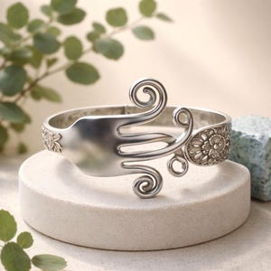 May include: Silver-toned metal bracelet featuring a fork design with decorative swirls and floral accents. The bracelet is displayed on a white circular stand, showcasing its unique craftsmanship and design. The bracelet's width is approximately 1 inch.
