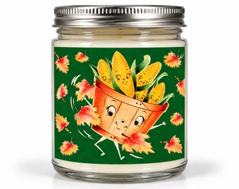 May include: A clear glass candle jar with a silver metal lid featuring a label with a dark green background. The label displays a cartoon wooden basket filled with smiling corn cobs, surrounded by scattered orange and red autumn maple leaves.