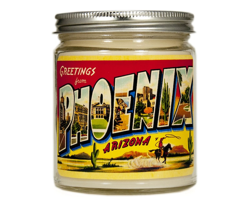 May include: A glass jar candle with a vintage postcard design featuring the text "Greetings from Phoenix, Arizona". The postcard image shows a cityscape, cacti, and a cowboy riding a horse.