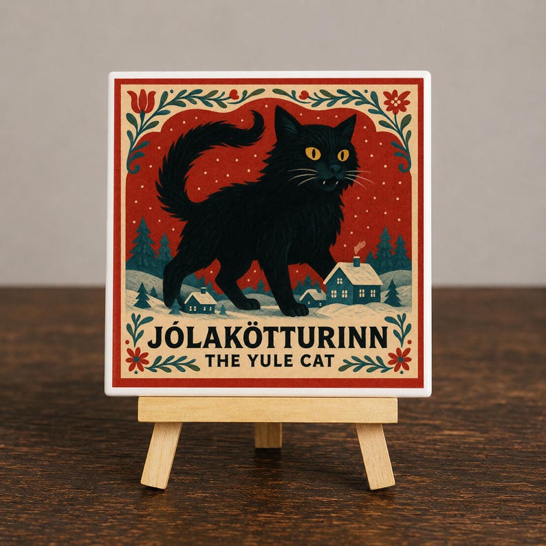 May include: A square ceramic tile featuring a black cat illustration on a red background with snow, pine trees, and small houses. The text reads J&Oacute;LAK&Ouml;TTURINN THE YULE CAT. The tile is displayed on a small wooden easel stand.