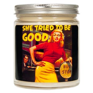 May include: A glass jar candle with a white lid and a vintage-style illustration of a woman in a red sweater and yellow skirt. The text "She Tried To Be Good" is printed in yellow on a black background.