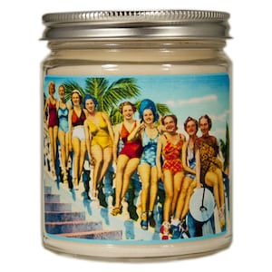 A glass jar candle with a silver lid. The label on the jar features a vintage illustration of women in swimsuits standing on a staircase. The background is a blue sky with palm trees.