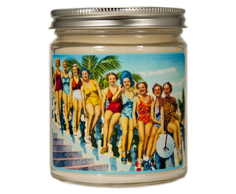 A glass jar candle with a silver lid. The label on the jar features a vintage illustration of women in swimsuits standing on a staircase. The background is a blue sky with palm trees.