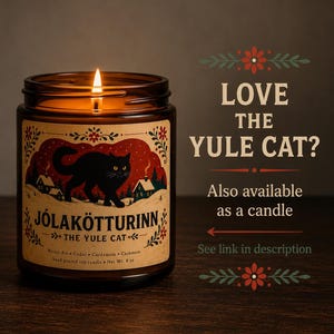 May include: A lit soy candle in an amber glass jar with a label featuring a black cat, snowy houses, and the text J&Oacute;LAK&Ouml;TTURINN THE YULE CAT. The label lists Winter Air, Cedar, Cardamom, and Cashmere scents with a net weight of 8 oz.