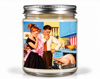 May include: A clear glass candle jar with a silver metal lid featuring a label with a vintage-style illustration of a person in a black dress and a person in a white shirt looking at a roasted turkey in an oven within a blue kitchen setting.