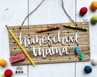 May include: A wooden sign with the words "homeschool mama" written in white letters. The sign is made from a ruler with a yellow pencil and a pink eraser on top. The sign is surrounded by colorful felt balls.