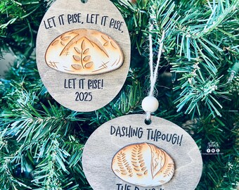 May include: Two wooden Christmas ornaments with bread illustrations and text. One says "LET IT PISE, LET IT PISE, LET IT PISE! 2025". The other says "DASHING THROUGH! THE DOUGH! 2025". Both have twine hangers.