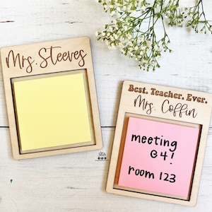 May include: Two wooden note holders with engraved names and space for sticky notes. The top holder says "Mrs. Steeves" and the bottom holder says "Best. Teacher. Ever. Mrs. Coffin". A pink sticky note in the bottom holder says "meeting 64! room 123".