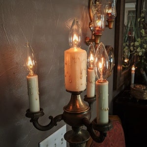 May include: A gold candelabra with four candle holders, each with a clear glass bulb and a white candle. The candles are dripping wax. The candelabra is on a wall with a light switch.
