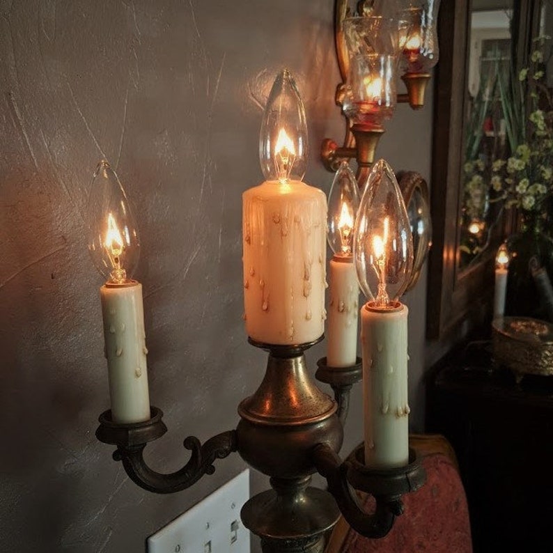 May include: A gold candelabra with four candle holders, each with a clear glass bulb and a white candle. The candles are dripping wax. The candelabra is on a wall with a light switch.