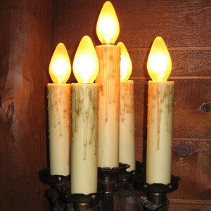 May include: A set of six candle-shaped light bulbs with a warm yellow glow, mounted on a dark brown metal candelabra. The bulbs are arranged in a circular pattern, with the center bulb slightly higher than the others.