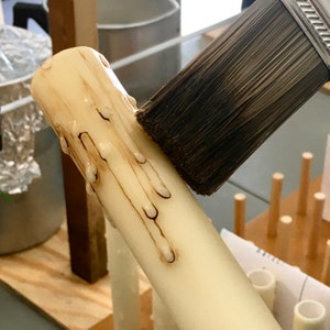 May include: A white candle with dripping wax is being brushed with a paintbrush. The candle is on a wooden surface.