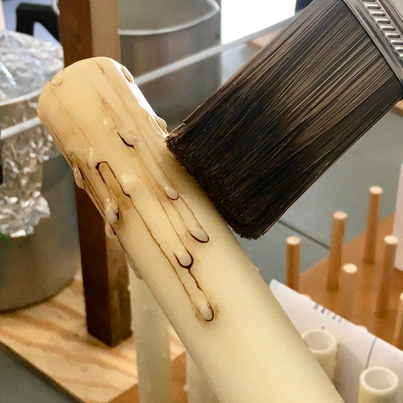 May include: A white candle with dripping wax is being brushed with a paintbrush. The candle is on a wooden surface.