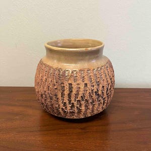 May include: A handcrafted, round ceramic vase with a textured, bark-like exterior. The vase has a wide opening and a light brown color, with a darker brown textured pattern on the body. The vase sits on a dark wooden surface.