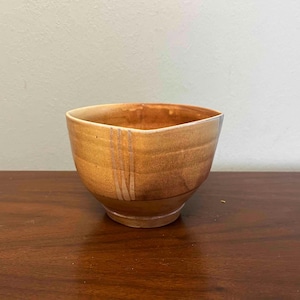 May include: A handmade ceramic bowl with a unique shape and a warm, earthy color palette. The bowl features a blend of brown and tan hues, with vertical stripe accents. The bowl is sitting on a wooden surface.