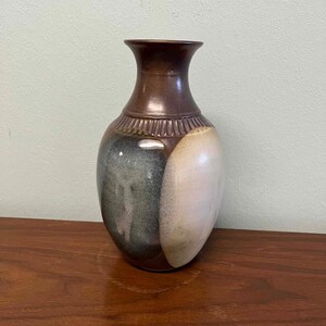 May include: A ceramic vase with a brown neck and rim, transitioning to a textured band, and a body divided into sections of green, white, and brown. The vase sits on a wooden surface.