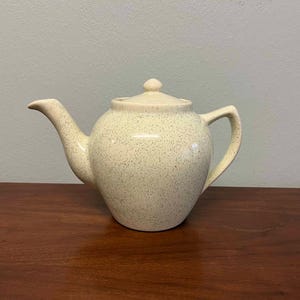 May include: A speckled cream teapot with a rounded body and a lid. The teapot has a spout and a handle.