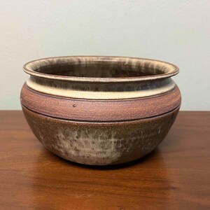 May include: A round ceramic bowl featuring a textured, earthy brown and tan glaze. The exterior displays a horizontal band of darker brown around the middle, transitioning into a lighter, mottled cream and brown finish toward the base and rim.