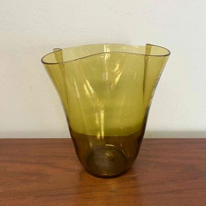 May include: A decorative, olive-green glass vase with a flared, wavy rim. The vase has a darker base and a lighter, translucent upper section. The vase is sitting on a wooden surface.