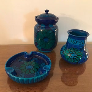 May include: A set of three blue and green ceramic pieces with floral designs. The set includes a lidded jar, a vase, and an ashtray.