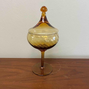May include: Amber-colored glass apothecary jar with a lid. The jar has a round base, a twisted stem, and a bulbous body. The lid is conical with a small knob. The glass has a warm, inviting hue, and the design is vintage.