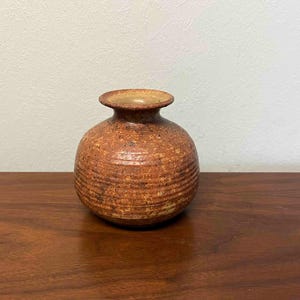 May include: A small, round, ceramic vase with a narrow neck and a wide, rounded body. The vase is a warm, earthy brown color with horizontal textured lines. The vase is sitting on a wooden surface.