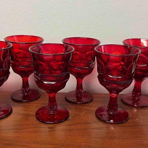 May include: A set of eight ruby red glass goblets featuring a textured, circular pattern on the bowl. Each goblet has a short, sturdy stem and a wide, circular base, providing a consistent look across the entire collection of drinking vessels.