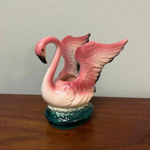 May include: A ceramic swan-shaped planter featuring a glossy finish with a white body, pink-tipped wings with black accents, and a dark green base molded to resemble foliage. The swan has a curved neck and an open cavity for holding items.