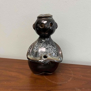 May include: A ceramic figurine with a dark brown glaze and a unique, stylized design. The figure has a rounded body, a small hat, and facial features including two eye holes and a small mouth. The glaze has a speckled, metallic appearance.