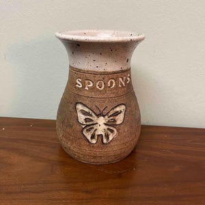 May include: A ceramic spoon holder with a brown base and a speckled off-white rim. The word "SPOONS" is embossed on the front, above a carved butterfly design. The holder is on a wooden surface.