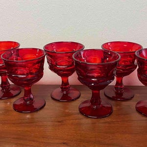 May include: A set of eight matching ruby red glass goblets, each featuring a short stem, a circular base, and a wide bowl with a textured, faceted surface pattern. The glassware is arranged in two rows, showcasing a deep, translucent red color.
