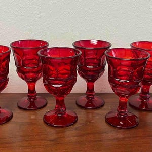 May include: A set of seven matching red glass goblets, each featuring a textured, dimpled pattern on the bowl, a short stem, and a circular base. The glassware is uniform in color and design, displaying a deep, translucent ruby red hue throughout.