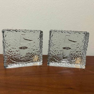 May include: Two clear, square glass candle holders with textured surfaces. Each holder features an etched design of a bird and a circular pattern. The candle holders are displayed on a wooden surface.
