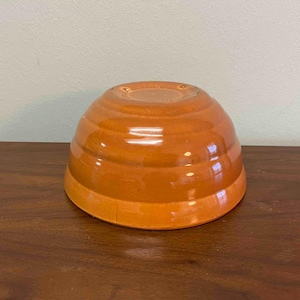 May include: A glazed, orange ceramic mixing bowl. The bowl has a rounded shape with horizontal ridges around the exterior. The bowl is sitting on a dark brown wooden surface.