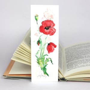 May include: Watercolor bookmark featuring a vibrant red poppy with green leaves and a bud. The floral design is painted on a white rectangular bookmark. The bookmark is placed between the pages of an open book.