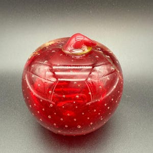 May include: A round, red glass decorative object with a small, curved handle on top. The glass has a bubbly texture and is a deep, transparent red color. The object is likely a decorative piece or a small container.