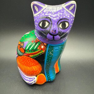May include: A vibrantly painted ceramic cat figurine. The cat features a purple head with white polka dots and large black eyes. Its body is adorned with colorful patterns in green, orange, and blue, showcasing a folk art style.