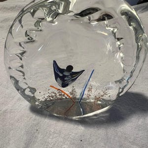 May include: A clear glass ashtray with a textured rim and a decorative design inside. The design includes a blue and black abstract shape, with orange and blue lines. The ashtray sits on a white surface.