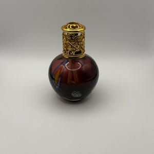 May include: A decorative glass bottle with a round shape and a gold-colored filigree top. The bottle has a dark burgundy base with colorful streaks of yellow, orange, and blue. The bottle is likely used for fragrance or essential oils.