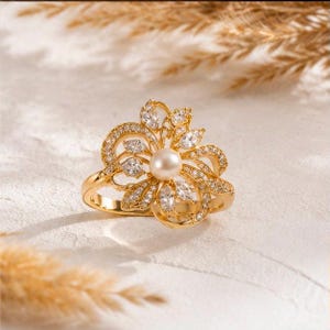May include: A gold ring featuring a central round white pearl surrounded by a floral-shaped arrangement of marquise-cut and round clear stones. The metal band and the petals are encrusted with small clear stones, creating a detailed design.