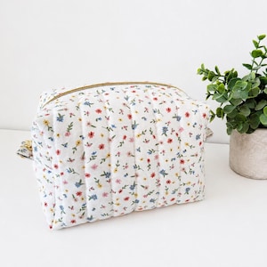 May include: A rectangular quilted pouch featuring a white fabric base with a small-scale floral pattern in red, yellow, and blue. The pouch has vertical stitching lines and a gold-toned zip closure along the top edge.