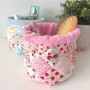 May include: Two fabric storage baskets. The front basket is pink with a strawberry pattern and a pink terry cloth lining. The second basket is blue with a patchwork design. The pink basket holds a hairbrush, perfume, and makeup.